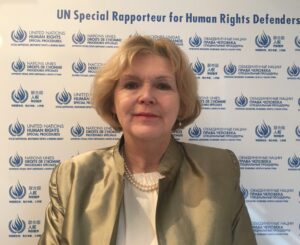 UN Special Rapporteur Mary Lawlor standing in front of a backdrop featuring the logo of the Special Procedures of the United Nations, at the top of which is written "UN Special for Human Rights Defenders". 