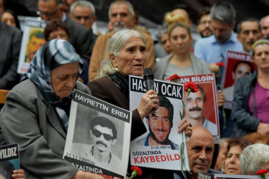 A crowd of women and men from the Saturday Mothers/People movement in Turkey are holding signs with photos of victims of enforced disappearances. An old woman is speaking with a microphone.