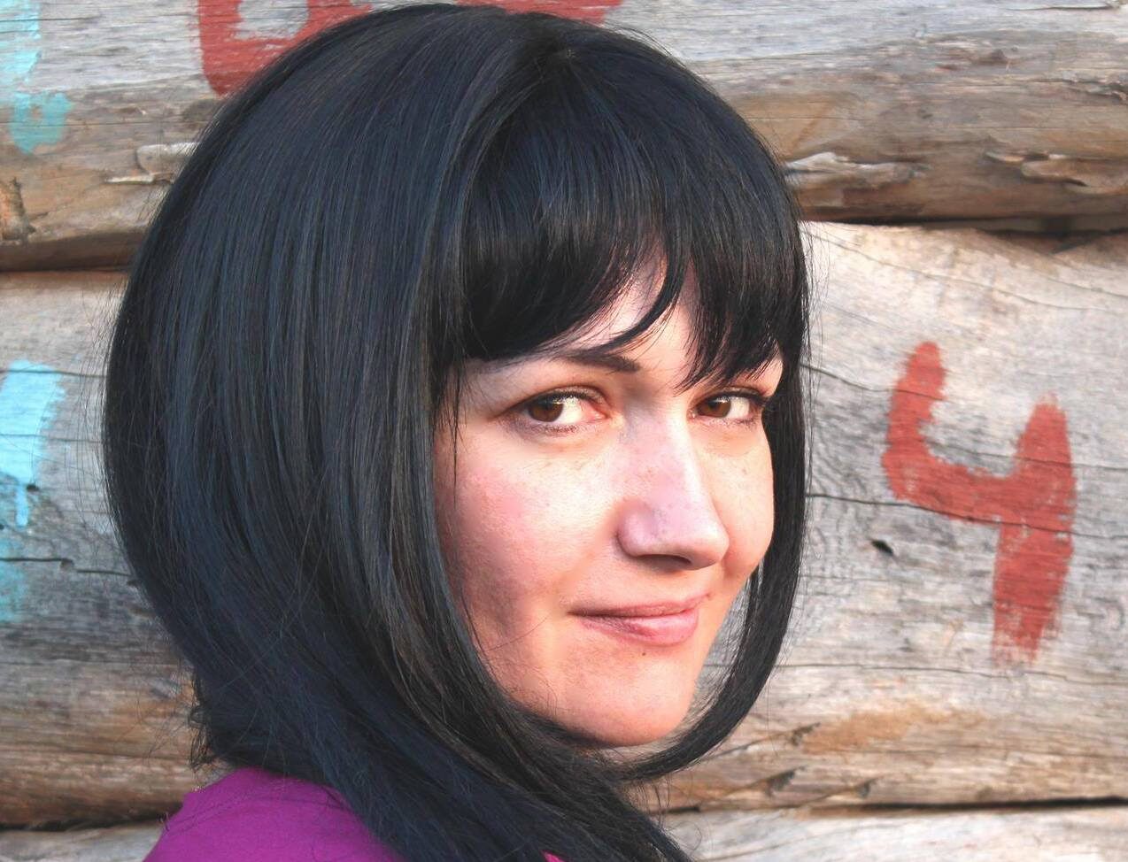 Irina Danilovich wears a purple t-shirt and looks at the camera. Her shoulders and head are visibile as she stands against a wall of wooden logs