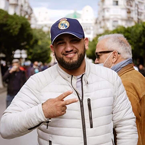 Picture of Zaki Hannache in the street, wearing a white coat and a baseball cap and making a peace sign with two fingers on his hand.