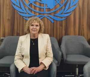 Mary Lawlor in a white jacket sits facing the camera in front of a blue UN logo