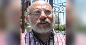A close-up portrait photo of Dr. Ahmed Amasha, outside on a sunny day. He is wearing glasses and donning a white beard. He is wearing a red and white checkered shirt.