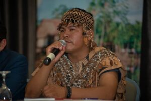 This image shows Herlín Odicio Estrella in traditional indigenous clothing, speaking into a microphone