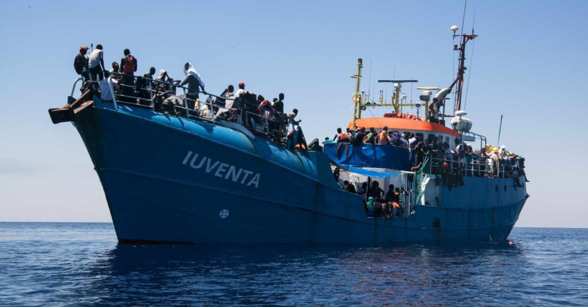 The image shows a blue boat where Iuventa is written in white letters. Many people are standing on the boat and clinging to its railing.