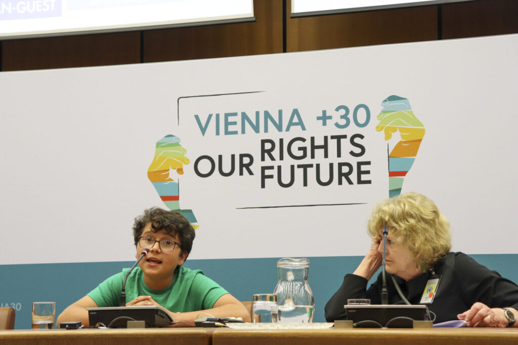 Francisco Vera Manzanares, a young human rights defender from Colombia, is speaking at a panel during the 2023 Vienna Youth & Child Human Rights Defenders Conference. He is wearing glasses and a green T-shirt. To his left is seated UN Special Rapporteur on Human Rights Defenders Mary Lawlor, who is looking at him while listening to his speech being simultaneously interpreted through an earphone. Behind them is a screen that reads Vienna + 30 Our Rights Our Future.  