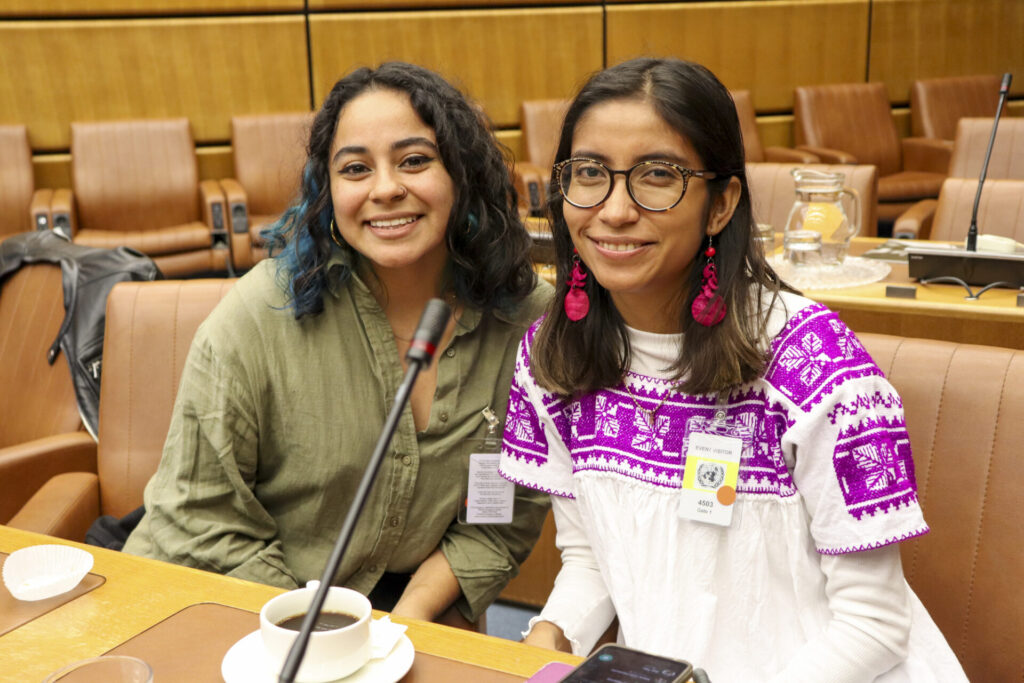 Young Human Rights Defenders Maria José Benavides from Honduras and Osmayra Solorio Loeza from Mexico are seating next to each other in the audience at the 2023 Vienna Youth & Child Human Rights Defenders Conference. They are smiling and looking at the camera. Maria José has black wavy hair with a strand of blue hair and is wearing a khaki shirt. Osmayra has dark brown hair and is wearing bright pink earrings with an embroidered white and purple blouse. 