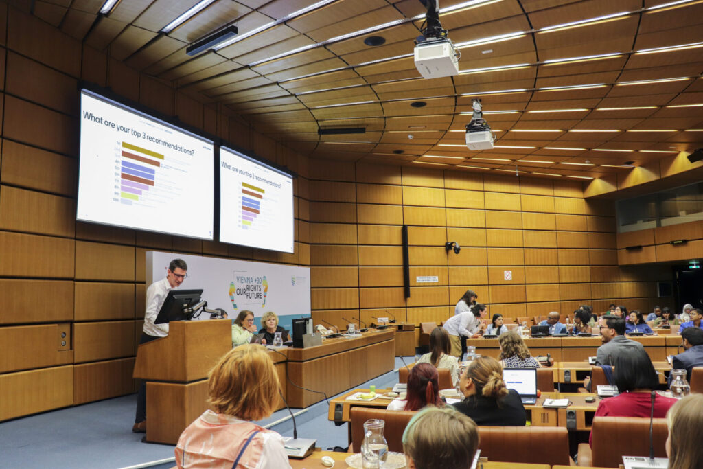A wide-angle picture of the room where the 2023 Vienna Youth & Child Human Rights Defenders Conference took place, at the UN Office in Vienna. Members of the UN Special Rapporteur on Human Rights Defenders’ team are on the stage, with one man standing at the lectern and reading from a monitor. Above them are screens showing the live results of a vote by the audience on recommendations for the protection of young human rights defenders. 