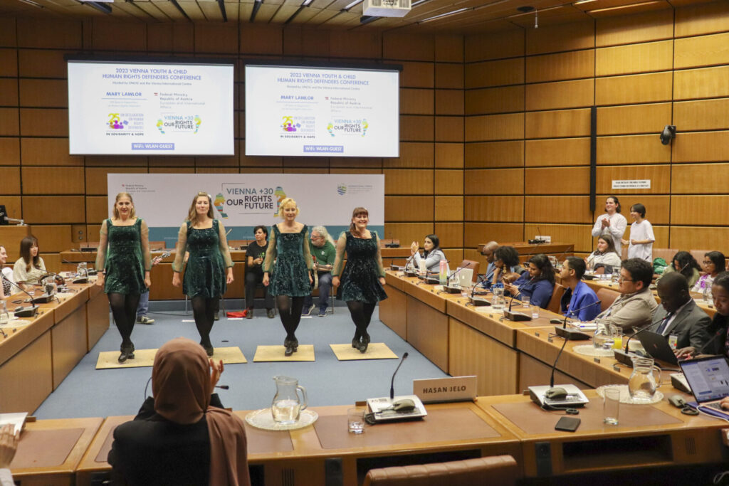 Four women wearing dark green dresses are performing an Irish traditional dance in the middle of the room where the 2023 Vienna Youth & Child Human Rights Defenders Conference took place. Behind them is a four-person band featuring guitars, a violin and drums. Participants of the conference are watching their performance, some clap while others film using their phone. 