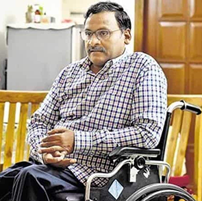 A photograph of GN Saibaba sitting in his wheelchair indoors, with a wooden door in the background