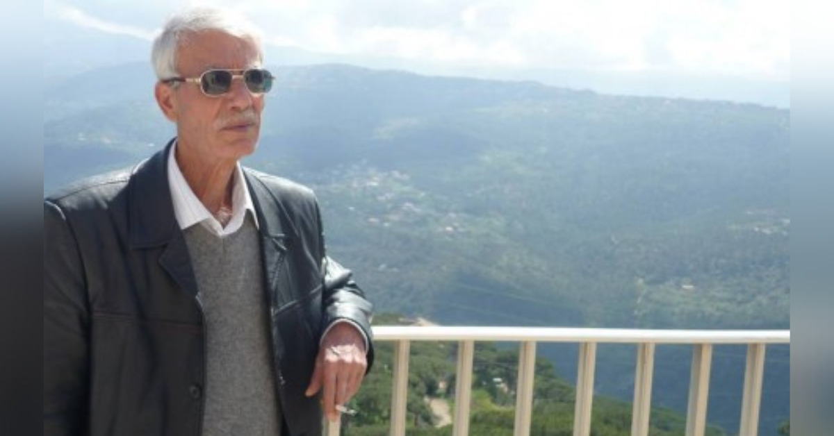 Syrian human rights defender Jdea Abdullah Nawfal is posing in front of a hilly landscape, his arm rested on a ledge and holding a cigarette. He has white hair and a moustache. He is wearing sunglasses and a black leather jacket.