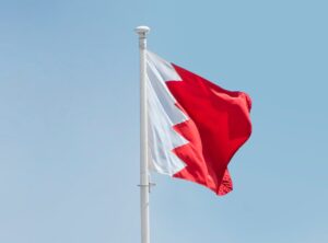 The flag of Bahrain waving at the top of a pole against a blue sky background.