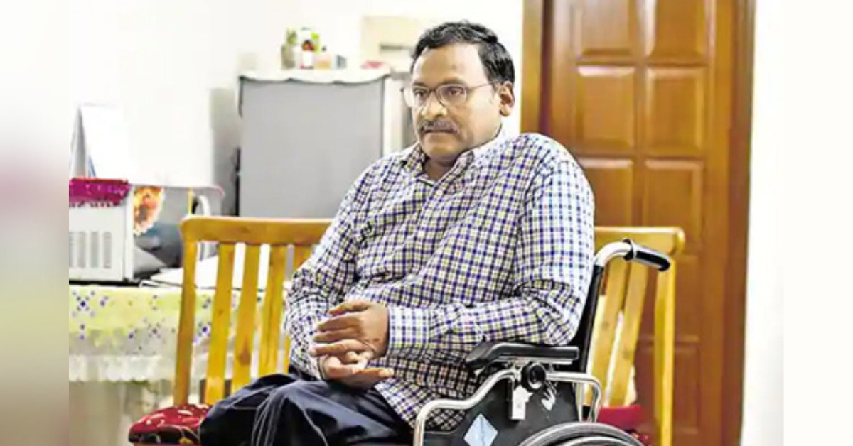 A photograph of GN Saibaba sitting in his wheelchair indoors, with a wooden door in the background.