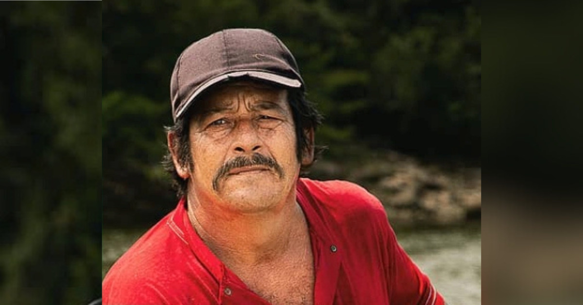 A portrait photograph of Colombian human rights defender Alirio Perdomo in an outdoor setting. He has black hair and a black moustache, and is wearing a black cap and a red tee-shirt.