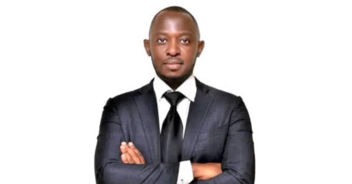 Bob Barigye is standing with his arms crossed on his chest. He is wearing a black formal suit in front of a white background.