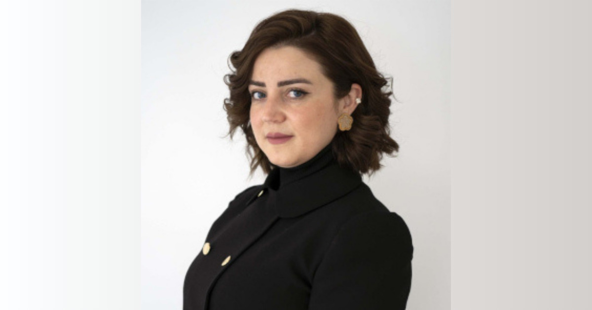 A portrait photograph of Syrian woman human rights defender Hiba Ezzideen Al-Hajji against a white background. She is wearing a black turtleneck and jacket, and has her brown wavy hair in a chin length bob.