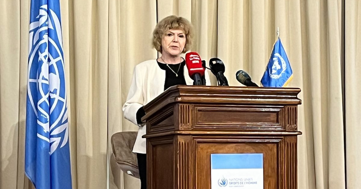 UN Special Rapporteur on Human Rights Defenders Mary Lawlor is speaking into microphones on a stage during the end of mission press conference in Algeria. Behind her is the flag of the United Nations.