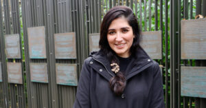 Poet and writer Nikita Gill is standing outdoors, in front of the Human Rights Defenders Memorial in Dublin. She is smiling at the camera. She has long black hair tied into a braid, a ring in her nose and is wearing a black jacket.