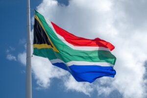 The flag of South Africa is flying in the wind. Behind it is the blue sky with a white cloud.