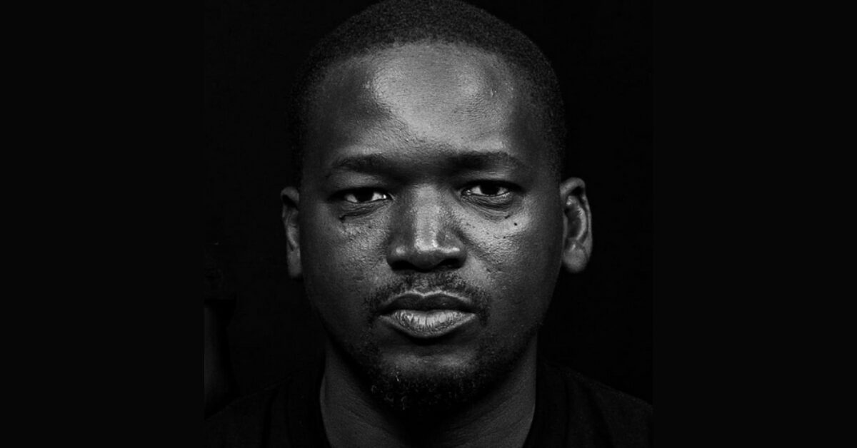 A black and white close-up shot of Senegalese human rights defender Aliou Sané looking straight at the camera against a black background.