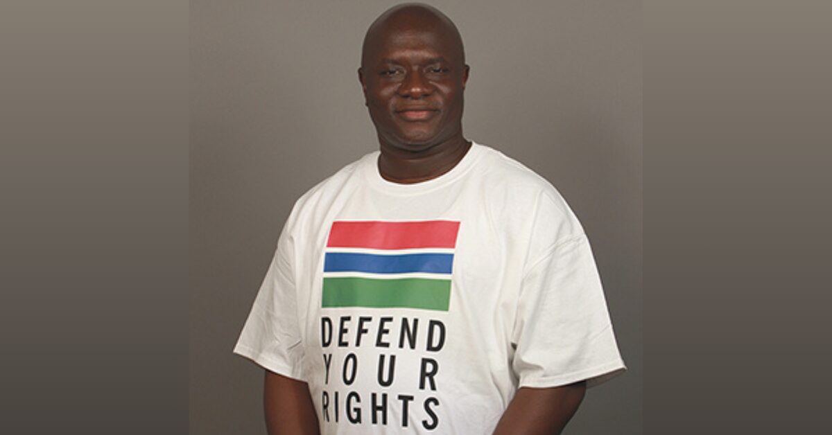 Gambian human rights defender Madi Jobarteh standing in front of a neutral background, wearing a white T-shirt with the flag of the Gambia, underneath which reads "Defend your rights".
