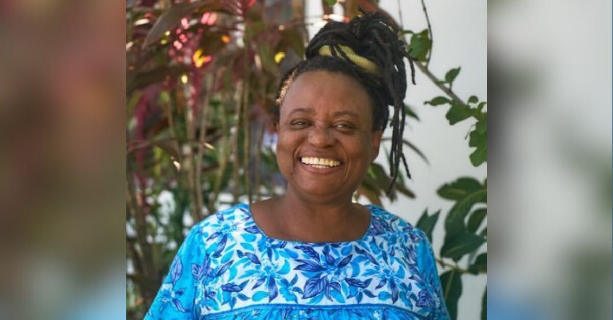Honduran woman human rights defender Miriam Miranda, a black woman, is smiling at the camera. Her hair is tied in an updo and she is wearing a bright blue top. In the background is some foliage.