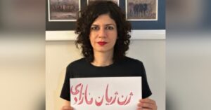 Iranian woman human right defender Nasim Soltanbeygi holding a sign with writing in Farsi in red. She has curly black shoulder-length hair and is wearing red lipstick and a black T-shirt.