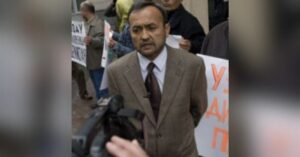 Human rights defender Bakhrom Khamroev is standing amongst protesters who are holding signs, and speaking to a journalist filming him. He is wearing a brown suit and tie.