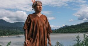 Peruvian human rights defender and indigenous Asháninka, Ángel Pedro Valerio, is standing in the sun outdoors, in front of a river in a lush mountain landscape. He is wearing a traditional headpiece with pearls and orange feathers and a large brown garment. His face is painted with red marks.