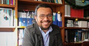 Colombian lawyer German Romero Sanchez sitting at a desk in front of a bookshelf containing files and books. He has long black hair tied into a ponytail, a short beard and wears glasses. He is smiling at the camera.
