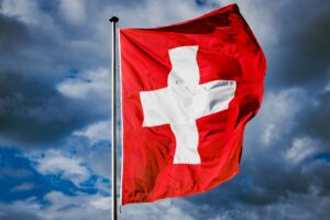 The flag of Switzerland flying in the wind under a blue sky with dark clouds.