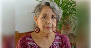 Mexican woman human rights defender Isela Gonzales Diaz is photographed in an indoor setting, sitting on a wooden chair with a plant in the background. She has short grey hair and is wearing a purple blouse with matching earrings.
