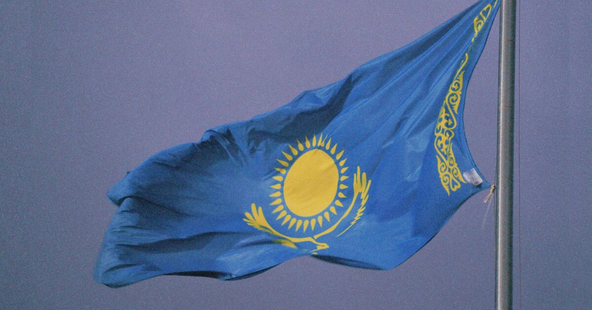 The flag of Kazakhstan blowing in the wind from the top of a flagpole, with a grey stormy sky in the background.