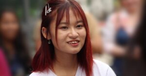 A young Thai woman, Anna Annanon, a human rights defender, is shown smiling. She has straight, shoulder-length red hair with a few hair clips, and is wearing a white shirt. Her earrings include a peace symbol, and the background is blurred with people in an outdoor setting.