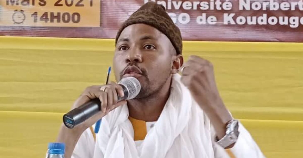 Burkina Faso human rights defender Daouda Diallo, wearing a traditional hat and white robe, is speaking into a microphone. He is seated in front of a yellow and red banner that partially reads 'Mars 2021' and 'Université Norbert ZONGO de Koudougou'.