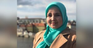 A woman, Lutfiye Zudiyeva, stands outdoors with a scenic cityscape in the background. She is wearing a teal hijab and a tan coat, smiling softly at the camera. The backdrop includes a river and various buildings under a cloudy sky.