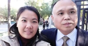 A selfie taken by Chinese woman human rights defender Xu Yan with her husband, human rights lawyer Yu Wensheng. Xu Yan is on the left, wearing a beige coat and smiling at the camera. Yu Wensheng is on the right, dressed in a suit and tie, with a serious expression. They are standing outdoors on a sunny day, with trees and a few people walking in the background.