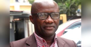 A portrait of Angolan lawyer Zola Ferreira Bambi, who is wearing glasses, a brown blazer, and a red and white checkered shirt. He is standing outdoors with blurred buildings and greenery in the background.