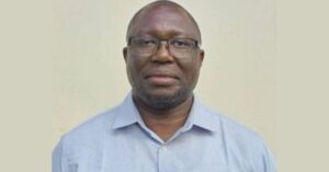 A portrait photograph of Kenyan human rights defender Chris Owalla, a black man with glasses wearing a light blue shirt, standing against a beige wall.