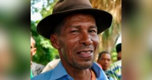 Colombian human rights defender Narciso Beleño, a middle-aged man with a friendly expression, is wearing a brown wide-brimmed hat and a blue shirt. His skin is sun-tanned, and his face shows wrinkles from a life spent outdoors. He is standing outdoors, with a blurred background of greenery and people.