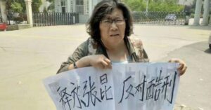 Undated photo of Chinese woman human rights defender Xu Qin holding a protest sign with handwritten Chinese characters. The sign appears to call for the release of someone and opposes torture, as she stands outside a building. Xu Qin has a serious expression, wearing glasses and a khaki jacket.