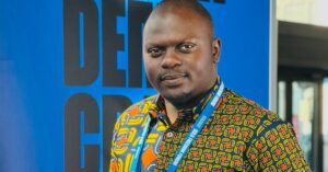 Portrait of Emmanuel Gumbo, a Zimbabwean lawyer and human rights defender, standing in front of a large blue banner. He is wearing a colorful, patterned shirt with orange, yellow, and blue accents, and has a conference lanyard around his neck. He is looking confidently at the camera.