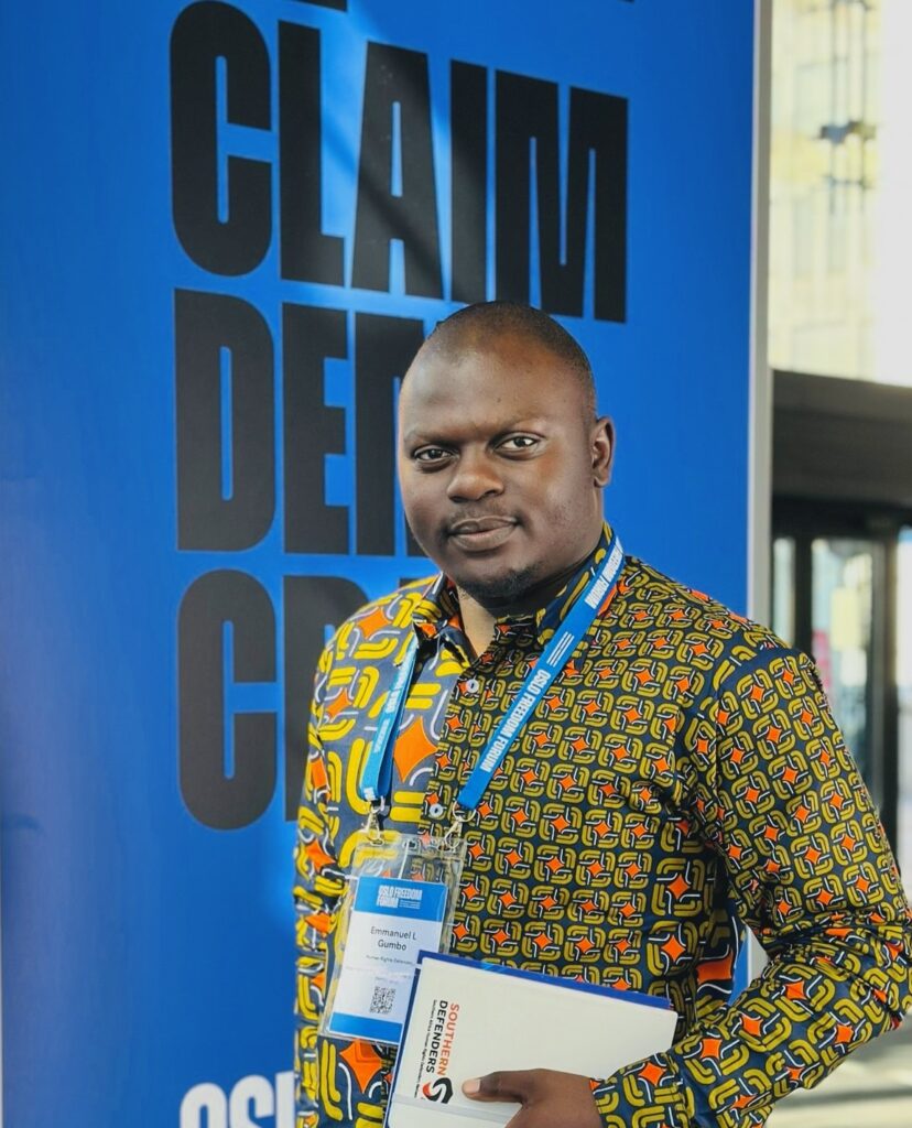 Portrait of Emmanuel Gumbo, a Zimbabwean lawyer and human rights defender, standing in front of a large blue banner with bold black text that reads 'CLAIM DEMOCRACY.' He is wearing a colorful, patterned shirt with orange, yellow, and blue accents, and has a conference lanyard around his neck with an ID badge. He is looking confidently at the camera.