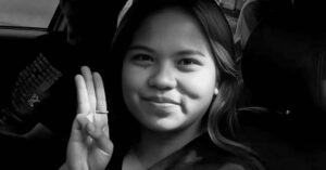 Black and white photograph of Thai activist Netiporn Sanesangkhom smiling at the camera and doing a three-finger salute, which is symbolically associated with the pro-democracy movement in Thailand and other countries in the region.