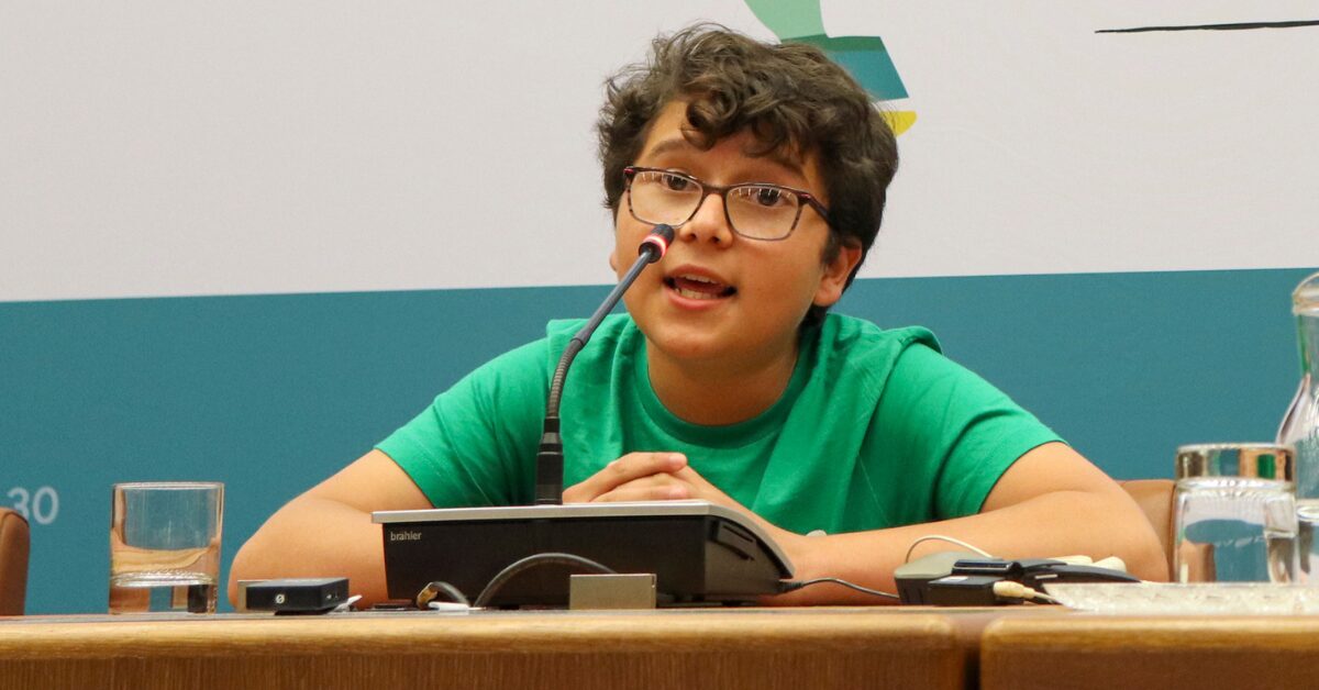 Young Colombian human rights defender Francisco Vera is speaking in front of a microphone during a conference. He has brown curly hair and is wearing glasses and a bright green tee-shirt.