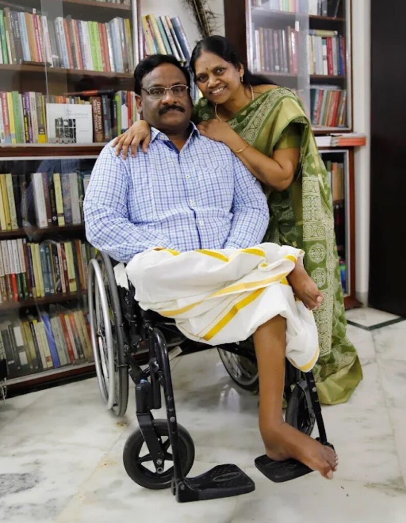 Photo of late Indian human rights defender GN Saibaba and his partner Vasantha. Saibaba, who uses a wheelchair, is seated wearing a checkered shirt and traditional white dhoti with a yellow border. Vasantha stands beside him, dressed in a green saree with intricate patterns, smiling warmly and resting her hands on his shoulders. They are in a cozy room filled with bookshelves that hold a variety of books and artwork.