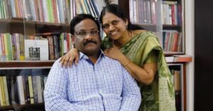 Cropped photo of late Indian human rights defender GN Saibaba and his partner Vasantha. Saibaba, who uses a wheelchair, is seated wearing a checkered shirt. Vasantha stands beside him, dressed in a green saree with intricate patterns, smiling warmly and resting her hands on his shoulders. They are in a cozy room filled with bookshelves that hold a variety of books and artwork.