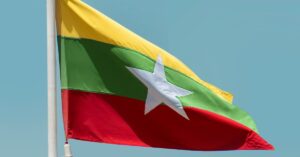 The flag of Myanmar, a white star over yellow, green and red horizontal stripes, flying from the top of a pole against a blue sky background.