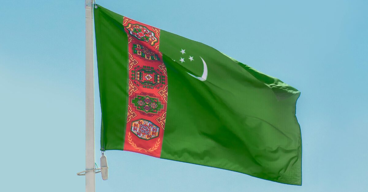 the flag of Turkmenistan flying in the wind from the top of a pole against a blue sky background.