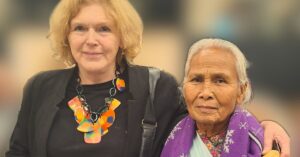 UN Special Rapporteur Mary Lawlor stands arm-in-arm with Nepali women's rights defender Rambati Chaudhari at an event. Mary, with shoulder-length blonde hair, wears a black outfit with a bold, colorful necklace. Rambati has silver hair tied back and wears a purple scarf with white snowflake patterns over traditional attire. Both women are smiling slightly. The background of the image is blurred.