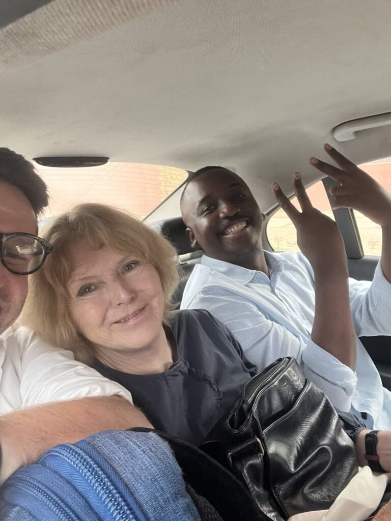 UN Special Rapporteur Mary Lawlor sitting in a car with her advisor Ed O'Donovan on the left (partially visible) and Nkanyiso from the Amnesty International regional section on the right. Mary smiles warmly at the camera, while Nkanyiso holds up a peace sign with a cheerful expression. The interior of the car and part of the outside scenery are visible through the windows.